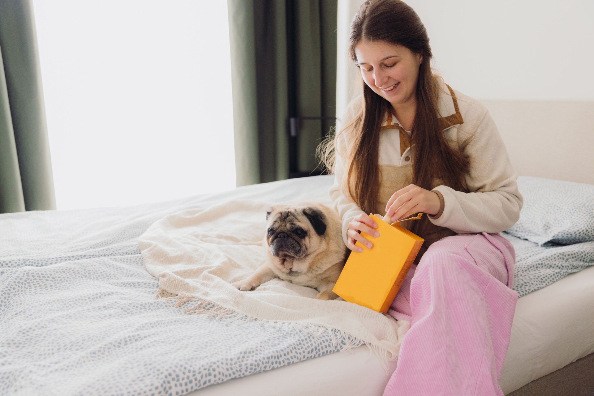 Happy Woman Feeding Her Cute Dog Snacks from a Box in Modern Bedroom